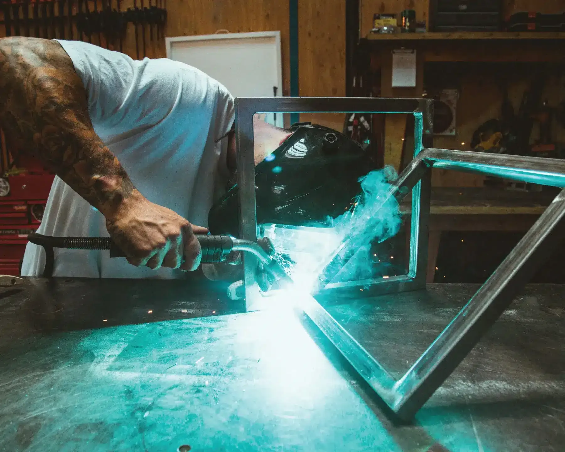 A production facility photograph showcasing in-house welding capabilities, with a welder creating a turquoise arc weld on a metal frame component for custom trade show exhibits. This image demonstrates Catapult3's skilled craftsmanship in structural fabrication and manufacturing quality.